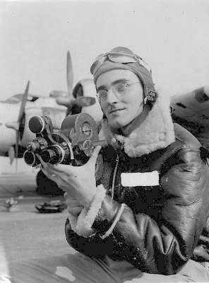 Close up view of Al Saldarini in wire-rimmed glasses wearing USAAF insulated flight jacker, and gunner's goggles atop a soft leather helmet. He's holding a 3-lensed camera. Wing and two engine cowls and propellers are visible above his shoulders.